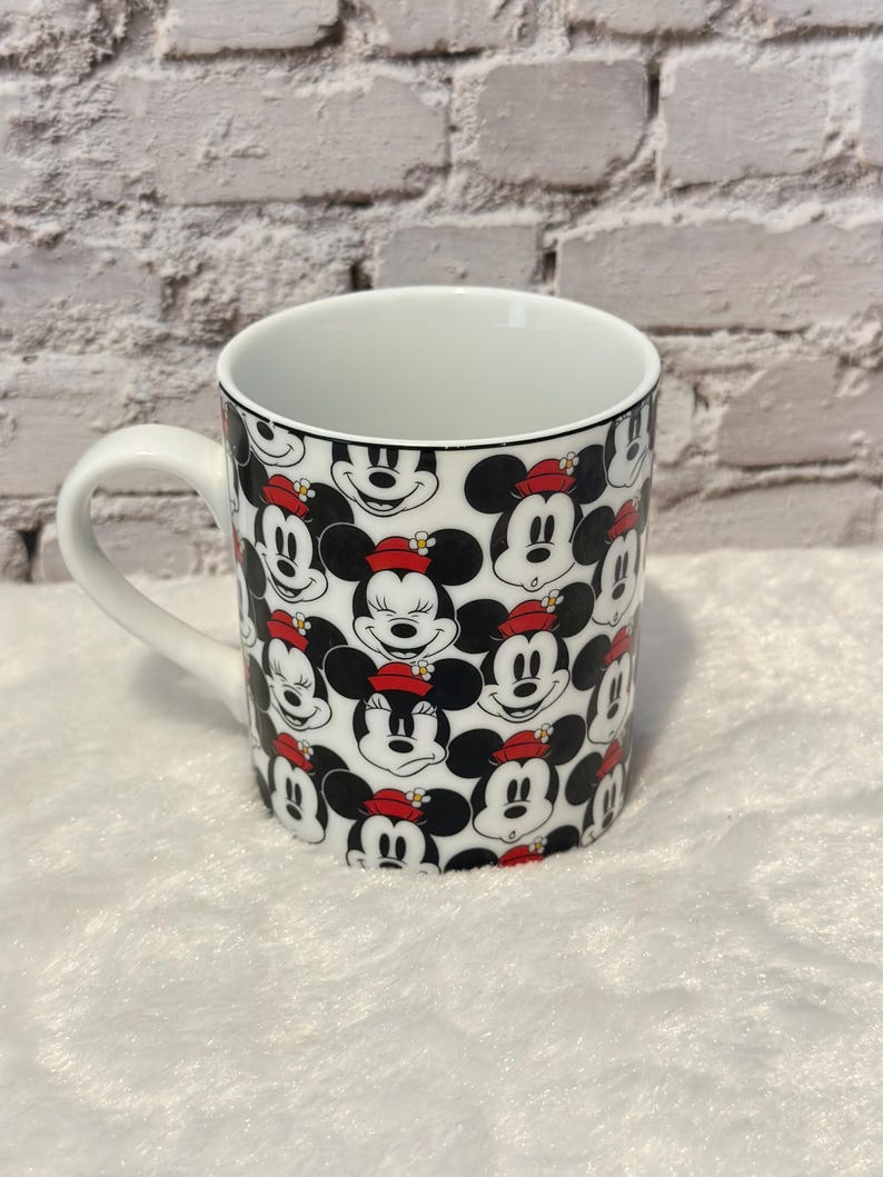 Puede incluir: Taza de cer&aacute;mica blanca con un patr&oacute;n repetido de caras de Mickey Mouse con sombreros rojos. La taza tiene un asa blanca y un borde negro. El fondo es una pared de ladrillos blancos y una superficie blanca y esponjosa.