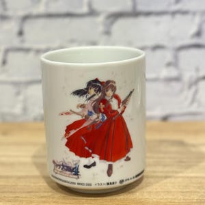 May include: White ceramic mug with a colourful anime illustration of two women in red and white dresses. The mug has a cylindrical shape and a smooth surface. The bottom of the mug has Japanese text.