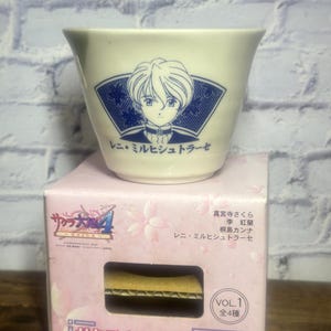 May include: A white ceramic cup featuring a blue and white illustration of a character from the "Sakura Wars" series. The cup rests on a pink box with Japanese text and the series logo, listing character names.
