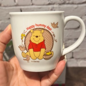 May include: White ceramic mug featuring Winnie the Pooh in a red shirt, with the text "Happy hummy days." The mug has a handle and is decorated with birds and a honey pot. A Disney product.