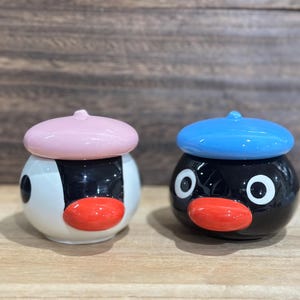May include: Two ceramic penguin-shaped containers. One is white and black with a pink beret and red beak. The other is black with a blue beret and red beak. Both have large white eyes and are on a wooden surface.