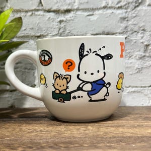 Pode incluir: Uma caneca de cerâmica branca com uma ilustração gráfica de um cão branco com uma camisa azul puxando um pequeno carrinho verde com um gato bege dentro. Acima do gato, há um balão laranja com um ponto de interrogação e o logótipo Fuwa Town.
