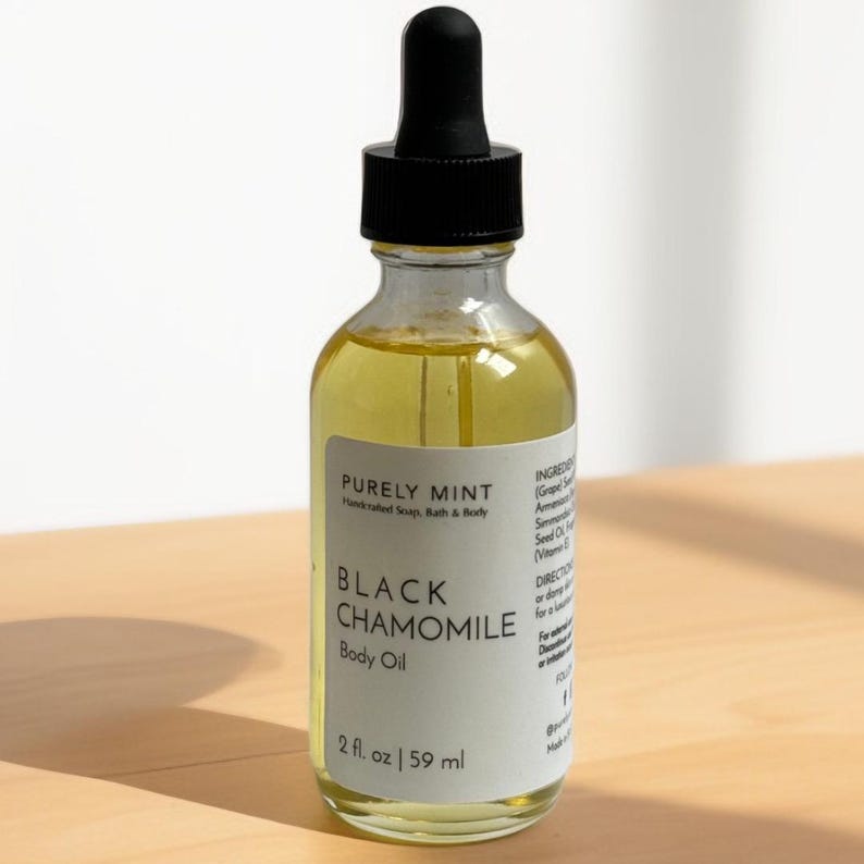 Black chamomile body oil in glass bottle, white label, black dropper top. Wood counter white background wall.