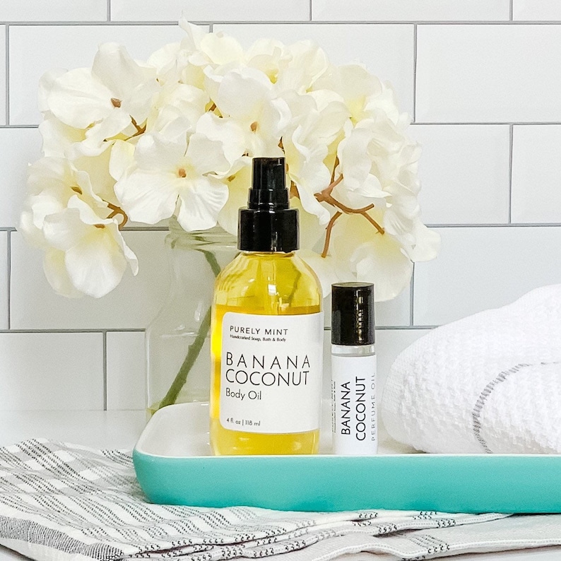 Organic Vegan Body Oil: Nut-Free, Sensitive Skin, Natural Moisturizer. Two bottles of banana coconut body oil, one in a clear glass bottle with a black cap and the other in a smaller roller bottle. The bottles are sitting on a teal tray with a white towel behind them.