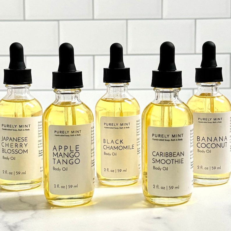 Organic Vegan Body Oil: Nut-Free, Sensitive Skin, Natural Moisturizer. Six glass bottles with dropper tops filled with yellow liquid. Each bottle has a white label with black text. The bottles are arranged in a row on a white tiled surface.