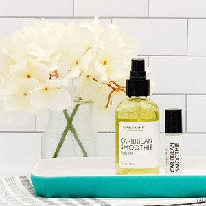 Two bottles of body oil with a white label that says 'Purely Mint' and 'Caribbean Smoothie Body Oil'. The smaller bottle is a roll-on and the larger bottle has a spray nozzle. The bottles are sitting on a teal tray. you
