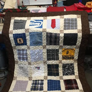 Könnte beinhalten: Eine braune gesteppte Decke mit braunem Rand. Die Decke besteht aus Quadraten aus verschiedenen Stoffen, darunter karierter Stoff, Denim und einfarbige Stoffe. Die Quadrate sind in einem Gittermuster angeordnet. Die Decke ist mit einem braunen Blumendesign verziert.