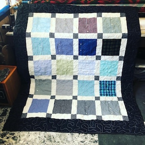 Könnte beinhalten: Eine Decke aus wiederverwendeten Hemden in verschiedenen blauen, grünen und weißen Karomustern. Die Decke ist in einem Rastermuster zusammengenäht, wobei schwarze Quadrate jedes Hemdquadrat trennen. Die Decke ist mit einem schwarzen Stoff mit einem dekorativen Wirbelmuster eingefasst.