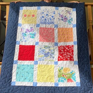 Könnte beinhalten: Eine blaue und weiße Patchwork-Decke mit verschiedenen Blumenmustern. Die Decke wird von einer jungen Person gehalten.