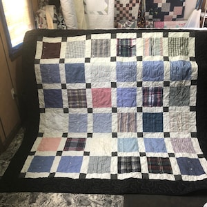 Pode incluir: Uma colcha de retalhos feita de tecido reutilizado, com uma variedade de padrões de xadrez e listras em tons de azul, branco e vermelho. A colcha é bordada com um tecido preto e tem um forro branco.
