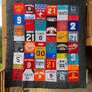 May include: A black and white quilt with a patchwork design made from different t-shirts. The quilt features various sports team logos, names, and numbers, including "Arsenal", "Madison County Colonels", "Da-Crack", "Suspects", "Lewis", "Durham", "Richmond All-Stars", "The Chosen One", "Celebrity Classic", "Home Lumber", "Flash", "Hawkes", and "Vivers".