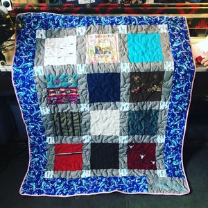 Könnte beinhalten: Eine blaue und weiße Steppdecke mit rosa Rand. Die Steppdecke besteht aus 12 Quadraten, jedes mit einem anderen Stoff. Die Quadrate sind in einem Gittermuster angeordnet. Die Steppdecke ist mit einem blauen und weißen Seepferdchenmuster verziert. Die Steppdecke hat den Text "To Grandma with Love" auf einem der Quadrate.