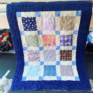 Könnte beinhalten: Eine blaue und weiße Patchwork-Decke mit einem blauen Rand. Die Decke besteht aus Quadraten aus verschiedenen Stoffen, darunter Blumenmuster, Karo und einfarbige Stoffe.