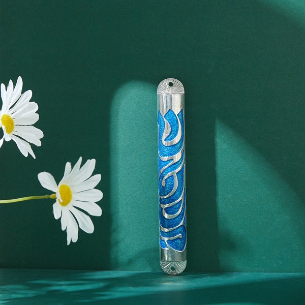 May include: A silver and blue mezuzah case with an ornate design. The case is cylindrical with a blue background and silver filigree-style patterns. The background is a deep teal green, with two white daisies with yellow centers.