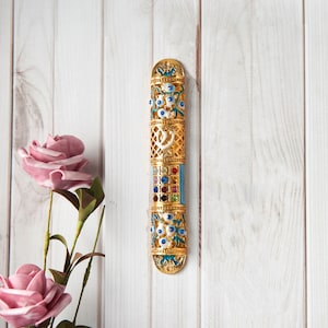 May include: A gold-colored mezuzah case with intricate floral and geometric designs. The case features blue, white, and colorful gemstone accents. The mezuzah is mounted on a white wooden surface, with pink roses in the background.