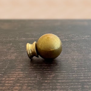 May include: A gold-toned metal finial with a round top and a smaller, cylindrical base. The finial is lying on a dark wood surface.