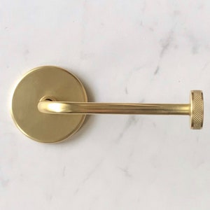 Op de afbeelding: Een gouden metalen handdoekhouder met een ronde basis en een knop aan het einde.
