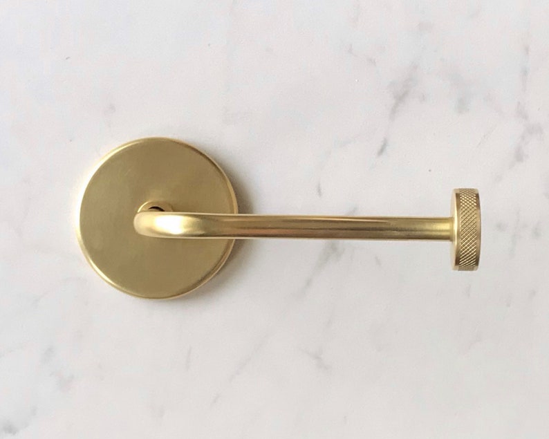 Op de afbeelding: Een gouden metalen handdoekhouder met een ronde basis en een knop aan het einde.