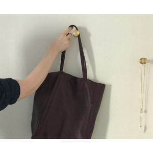 May include: Two gold-toned hooks mounted on a white wall. A brown leather tote bag hangs from one hook, and a gold chain necklace hangs from the other.