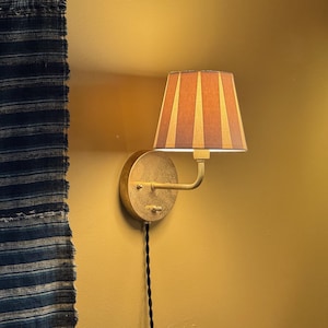 May include: A gold-toned wall sconce with a striped lampshade. The lampshade has a warm, orange and gold color scheme. The sconce is mounted on a mustard-yellow wall, with a black cord hanging down. A blue and white striped textile is visible on the left.