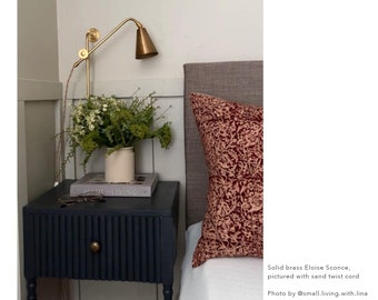May include: A dark blue nightstand with a fluted front and a brass knob. A book titled "Made for Living" is on top of the nightstand, along with a pair of glasses. A brass wall sconce with a cone-shaped shade is mounted on the wall behind the nightstand. A bouquet of white and green flowers in a white ceramic vase is on the nightstand. A red and white patterned pillow is on a bed behind the nightstand.