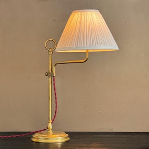May include: A brass desk lamp with a pleated white lampshade. The lamp has a curved arm and a red fabric-covered cord. The base is round and the lamp is on a dark surface.