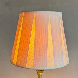 May include: A lampshade with a striped pattern in shades of beige and yellow. The lampshade is made of fabric and has a pleated design.