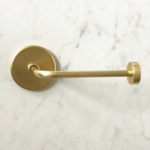 Op de afbeelding: Een gouden metalen toiletpapierhouder met een ronde basis en een gebogen arm. De houder is gemonteerd aan een witte muur.