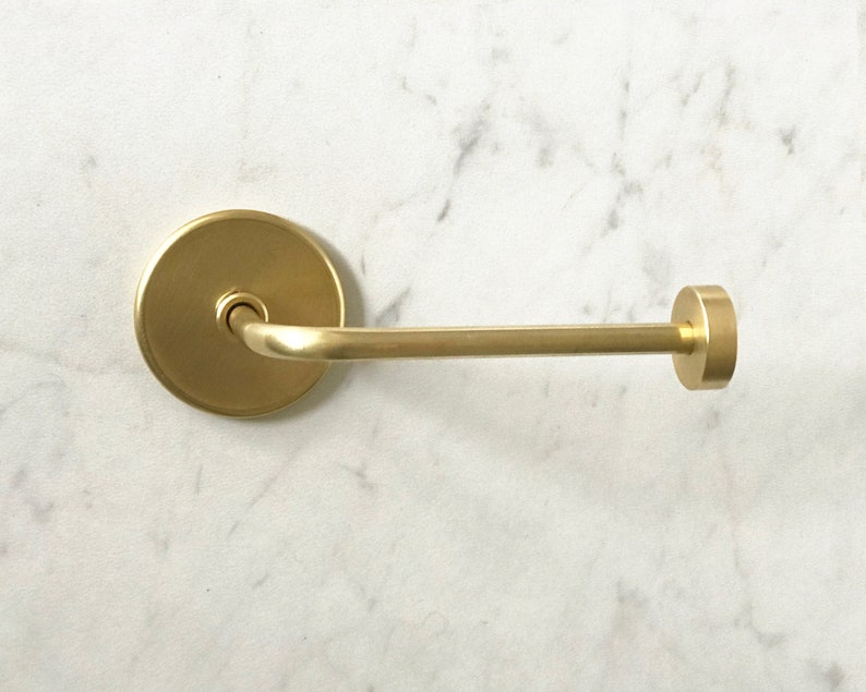 Op de afbeelding: Een gouden metalen toiletpapierhouder met een ronde basis en een gebogen arm. De houder is gemonteerd aan een witte muur.
