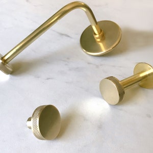 Op de afbeelding: Drie gouden metalen haken met een geribbeld ontwerp. De haken zijn van verschillende groottes en vormen. De grootste haak is een gebogen wandbevestiging met een ronde basis. De andere twee haken zijn kleiner en hebben een ronde basis met een korte steel.