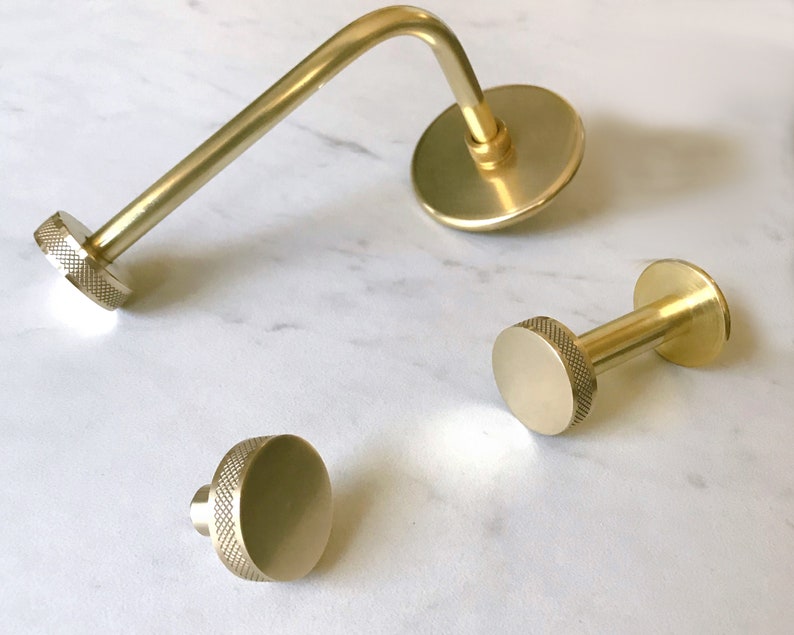 Op de afbeelding: Drie gouden metalen haken met een geribbeld ontwerp. De haken zijn van verschillende groottes en vormen. De grootste haak is een gebogen wandbevestiging met een ronde basis. De andere twee haken zijn kleiner en hebben een ronde basis met een korte steel.
