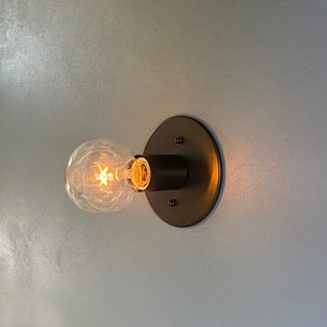 May include: A close-up of a wall-mounted light fixture with a round, brushed brass base and a single light bulb. The light bulb is turned on and emits a warm, yellow glow.