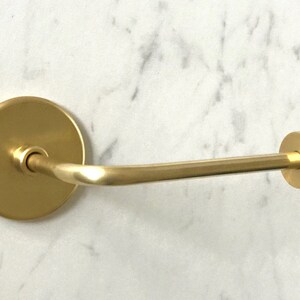 Op de afbeelding: Een gouden toiletpapierhouder met een ronde basis en een gebogen arm. De houder is gemonteerd op een witte marmeren muur.