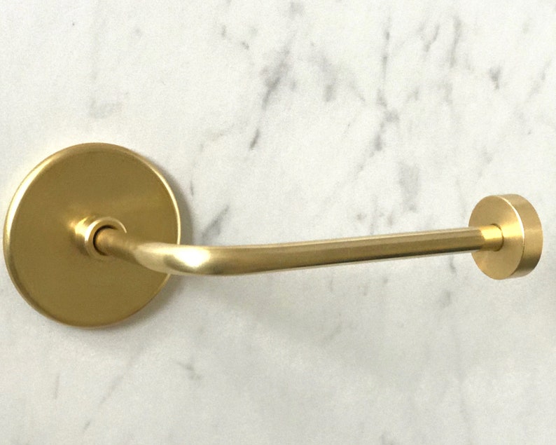 Op de afbeelding: Een gouden toiletpapierhouder met een ronde basis en een gebogen arm. De houder is gemonteerd op een witte marmeren muur.