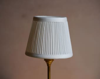 May include: A white pleated lampshade with a tapered, conical shape. The shade features vertical folds throughout its surface and is mounted on a metallic gold-toned base that extends downward from the center of the bottom opening of the shade.