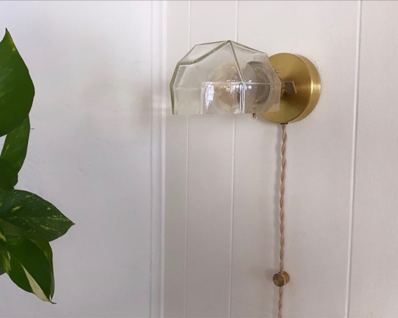 May include: A gold wall sconce with a clear glass shade and a brown cord. The sconce is mounted on a white wall.