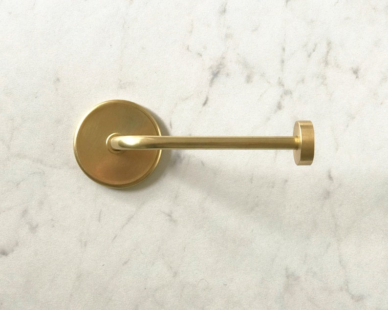 Op de afbeelding: Een gouden metalen toiletpapierhouder met een ronde basis en een gebogen arm. De houder is gemonteerd op een witte marmeren muur.