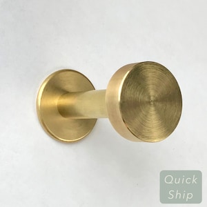 May include: A gold metal hook with a round base and a circular top. The hook is mounted on a white wall.