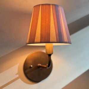 May include: A wall-mounted lamp with a striped fabric shade in shades of brown and beige. The lamp is mounted on a brass-coloured base.