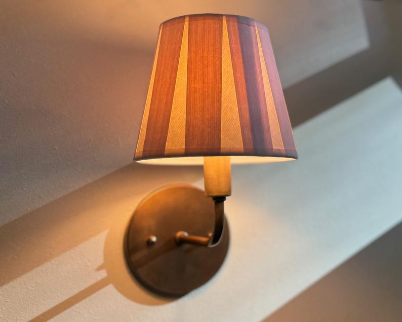 May include: A wall-mounted lamp with a striped fabric shade in shades of brown and beige. The lamp is mounted on a brass-coloured base.