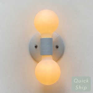 May include: White ceramic wall sconce with two frosted glass light bulbs. The sconce has a simple design with a rectangular base and two round bulbs.