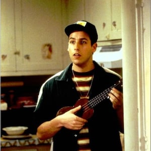 May include: A man wearing a black shirt with a striped undershirt and a black baseball cap is holding a brown ukulele. He is standing in a kitchen with white cabinets and a white tiled backsplash.