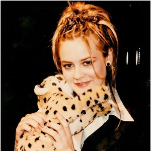 May include: A plush cheetah toy with a tan and black spotted pattern. The toy is held by a person wearing a black blazer and a white collared shirt. The background is dark.