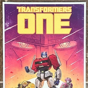 May include: A comic book cover featuring the Transformers. The title "TRANSFORMERS ONE" is in yellow block letters. Optimus Prime, Bumblebee, and other Autobots stand on a purple mountain against a red and orange background. "Limited Edition 2024" is in the lower left corner.