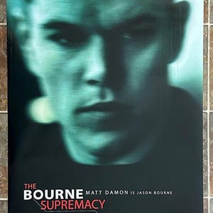 May include: Movie poster for "The Bourne Supremacy" featuring a close-up of Matt Damon's face in shades of teal and black. The title and release date, July 23, 2004, are displayed at the bottom, with the tagline "THEY SHOULD HAVE LEFT HIM ALONE" at the top.