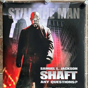 May include: Movie poster for the film "Shaft" starring Samuel L. Jackson. The poster features a black and white image of Jackson in a black leather jacket and jeans, walking down a city street. The title of the film is in large, white letters, and the tagline "Any Questions?" is below it. The poster is set against a red and orange background.