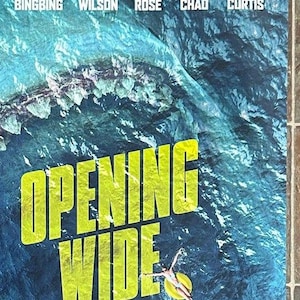 Puede incluir: Un póster de película para la película "The Meg" que presenta la boca de un tiburón gigante abierta de par en par con una persona en una balsa salvavidas nadando en el agua. El póster tiene el texto "Opening Wide" y "August 10 Only in Theaters" en letras amarillas. El póster también presenta los nombres de los actores de la película, incluidos Jason Statham, Rainn Wilson, Ruby Rose, Winston Chao y Cliff Curtis.