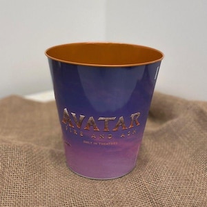 May include: A tapered metal bucket with a smooth finish, featuring a gradient design transitioning from deep purple at the top to a lighter pink at the base. The text AVATAR FIRE AND ASH ONLY IN THEATRES is printed in gold on the front surface.