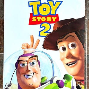 May include: A movie poster for Disney Pixar's "Toy Story 2" featuring Buzz Lightyear and Woody. The title is in yellow and red, with the film's stars' names at the top. The tagline "LIGHT THE TOYS ARE BACK!" is at the bottom.