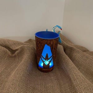 May include: A cylindrical cup with a brown, textured exterior resembling tree bark and a bright blue interior. A glowing blue triangular cutout on the front displays a stylized symbol. A small blue and white creature figure is attached to the rim.